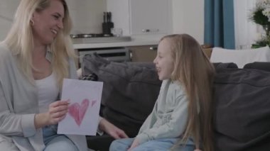 cheerful girl daughter gives greeting card with painted heart shape to mother as surprise present on happy Mothers day. mom and kid hugging and smiling happy family day