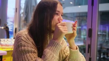 Süveter giyen, kahve içen, gülümseyen ve sonbahar kahve molasında pencereden dışarı bakan genç ve güzel bir kadın.