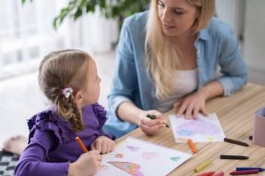 pre school art class for creativity development. teacher helps to little girl to learn drawing and painting on paper with colorful crayons.