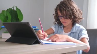 Kıvırcık saçlı, gözlüklü, uzaktan kumandalı, sevimli okul çocuğu. Dijital tablet kullanan konsantre çocuk, video dersinde öğreniyor..