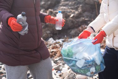 İki kadın plajda poşetlerde plastik çöp topluyor. Kirlilik felaketinin etrafına çöp döktü. Boş plastik şişeler ve kağıt atıkları