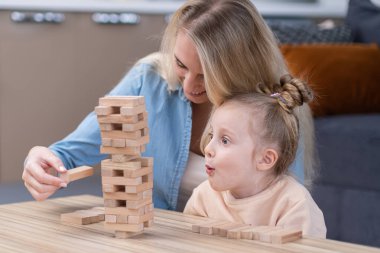 little daugher looking excited to Jenga wooden bricks tower, mother teaching cute girl to play board games, mom and kid spending time at home playing