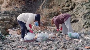 Sorumlu vatandaş çevreye zarar gelmesin diye plastikleri deniz kıyısından kaldırıyor. Eldivenli kadınlar plastik torbalara çöp topluyor. 