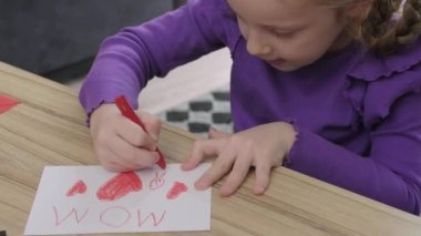 Little girl preparing a greeting card to mom with red heart painted and drawing MOM letters as gift for mothers day. colored pencils and crayon on table. 