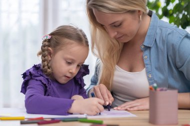Anne, kızına ödevlerinde yardım ediyor, okul ödevlerinde birlikte çalışıyor masa başında oturup renkli kalemlerle resim çiziyorlar..