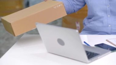 Kadın, ürün siparişlerini kontrol etmek için dizüstü bilgisayar ve telefon kullanıyor ve ekspres nakliye hizmetleri kullanarak çevrimiçi işini bir depodan yönetirken verileri kaydediyor.