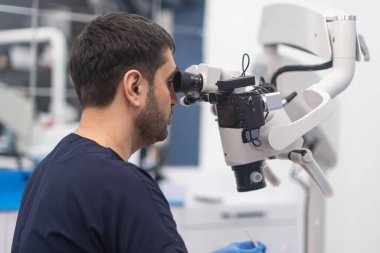 Modern diş mikroskobuna bakan sakallı doktor, diş hekimliği kliniğinde yenilikçi teknoloji kullanarak kanepe odaklı dişçide hastanın boşluğunu kontrol ediyor.