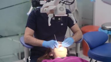 Modern klinik asistanı üniformalı diş mikroskobuyla dişlerini kontrol eden dişçi, kadın hastaların profesyonel muayenesinde doktora yardım ediyor.