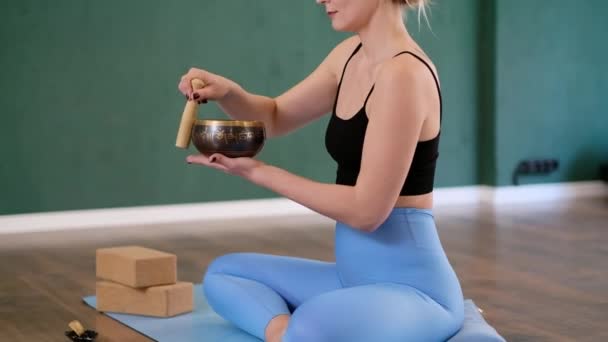 在工作室的职业瑜伽课上 一个女人坐在瑜伽旁边的蓝色垫子上 用西藏的歌唱碗来做冥想 会阻碍年轻女子尝试新的仪式 — 图库视频影像