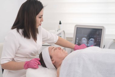 Kadın, profesyonel yüz bakımı görüyor ve lazer makinesiyle ultrason çekiyor. Doktor, ameliyat için cihaz ekranını kullanıyor. 