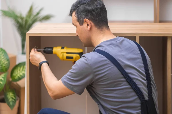 Wooden furniture industry man attaching door hinge to wooden cabinet side with screwdriver firm worker in overalls assembling new furniture for living room interior