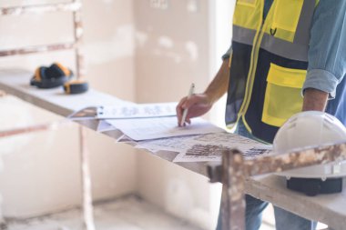 Profesyonel üniformalı işçiler inşaat belgelerini düzenliyor ve düzeltiyorlar. Dairenin güvenlik ekipmanları yenileme sürecinde odada duruyorlar.