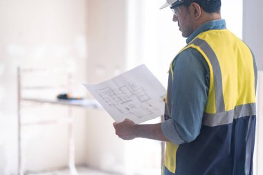 Tadilat firması çalışanı profesyonel üniformalı daire çizim uzmanı bitmemiş yatak odasındaki inşaat güvenlik ekipmanlarına karşı duruyor.