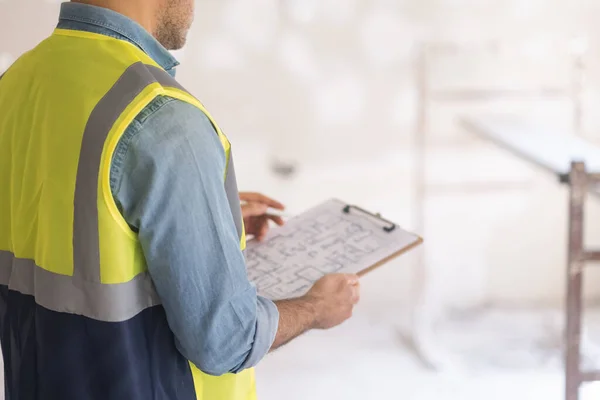 Yelek giyen ustabaşı büyük evin çizim planını kontrol ediyor. Pano ve kalem ellerinde. Profesyonel üniformalı adam odada yenileme işlemine hazırlanıyor.