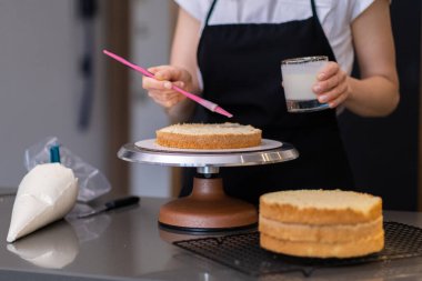 Yetenekli ev hanımı, yemek fırçasıyla taze pasta tabakalarını siyah önlüklü leziz kremalı kadınla süpürüp düğün partisi için mutfak masasında duran özel şekerleme yapıyor.