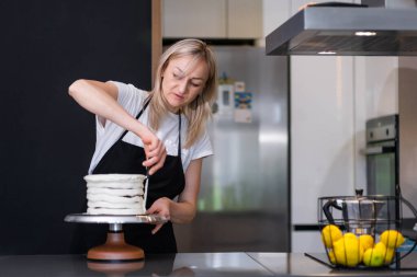 Yoğunlaşmış siyah önlüklü sarışın kadın, yuvarlak pastanın etrafına beyaz mastik serpiştirmekle meşgul. Modern mutfak pastacısının önünde durmuş, evde şekersiz pastanın tadını çıkartıyor.