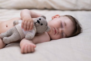 Sevimli küçük çocuk, elinde oyuncak ayıyla yumuşak yatağa uzanmış beyaz çarşaflı, tombul bir bebekle sabahları yatak odasında uyumaktan zevk alıyor.