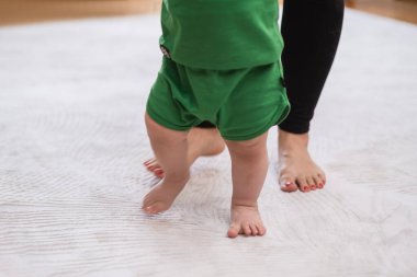 Tatlı yalınayak çocuk, anne meraklısı çocuğun elini tutarak beyaz halıda yürüyor. Annesinin büyümesi ve gelişimi sayesinde ilk adımları atmayı öğreniyor.