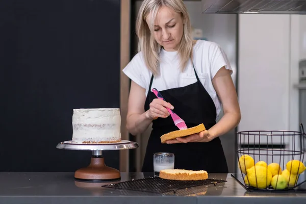 Focused blonde confectioner spreading white cream on cake layers with cooking brush standing at kitchen table woman in black apron making creamy pastry from organic products at home