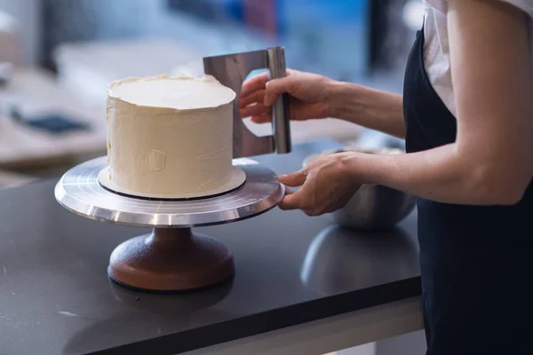 stock image Housewife in black apron processing white mastic on organic lactose free cake with pastry scraper standing at table woman using kitchen utensils to make delicious confectionery at home