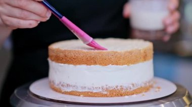 Siyah önlüklü sarışın kadın taze pasta tabaklarını kremayla süpürüyor. Pembe yemek fırçası kullanıyor. Mutfak masasında durmuş düğün pastasını hazırlıyor.