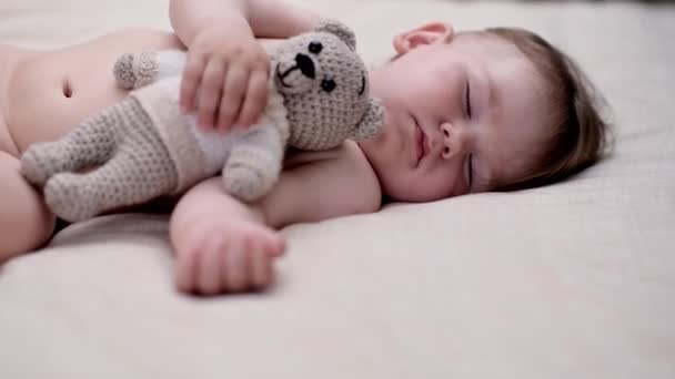 可愛いです男の子抱擁ニットクマおもちゃとともに手嘘柔らかいベッドの上に白いシートかわいいですぽっちゃり赤ちゃん楽しみますベッドルームで自宅で朝 — ストック動画