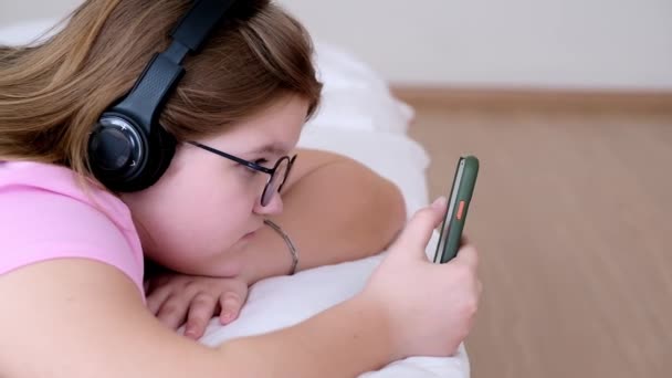 在网上冲浪时 在无线耳机里放轻松的青少年一边听讲座 一边戴着眼镜 手里拿着手机躺在床上 — 图库视频影像