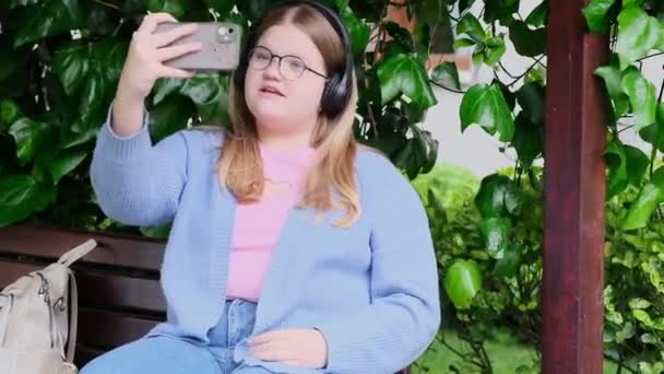 ヘッドフォンで太りすぎの10代の女の子は 現代のスマートフォンのブロンドプラスサイズの女子高生のカメラで自撮りは春の公園でベンチに座ってソーシャルメディアのための写真を撮ります — ストック動画