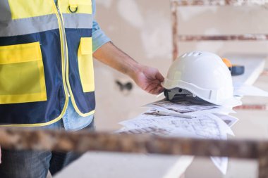 Yelekli mimar, inşaat sahasının güvenlik kuralları ve ekipmanlarında üniformalı adamın yenileme işlemine devam etmek için proje kağıtlarının yığınından beyaz miğfer alıyor.