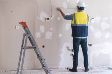 Profesyonel üniformalı, merdivenin yanında duran yetenekli bir işçi, oturma odasına soket yerleştirmeye hazırlanan, koruyucu kasklı bir adamla eski püskü bir duvarı ölçer.