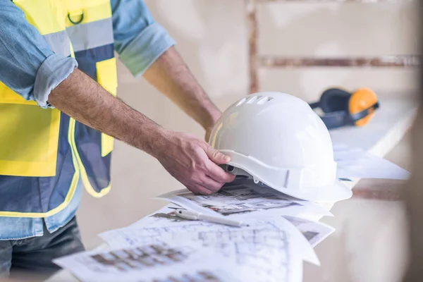 Skilled worker in uniform taking white protective helmet lying on heap of project papers specialist getting ready for renovation in room construction safety rules and equipment
