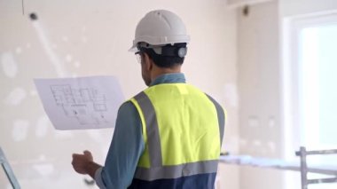 Koruyucu başlık okuma projesi çizimi işçisi inşaat güvenlik ekipmanları yanında duruyor. Üniforma konusunda uzman, ofis yenileme sürecine hazırlanıyor.