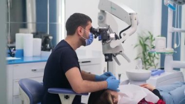 Koruyucu maskeli ve eldivenli yetenekli dişçi diş hekimliği kliniğinde çalışan diş mikroskobu uzmanı bir kadının dişlerini tedavi ediyor.