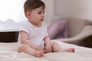 Beyaz takım elbiseli, tombul, tatlı bir bebek. Yumuşak yatakta oturmuş, sabah uyandıktan sonra yatak odasında dinlenen, yalınayak, sevimli bir çocuğa bakıyor.