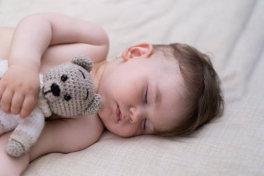 Siyah saçlı tatlı çocuk beşikte beyaz bir çarşafın üzerinde elinde ayı oyuncağı, elinde elleri ve gözleri kapalı masum tombul bebek sabahın erken saatlerinde bebek odasında uyuyor.