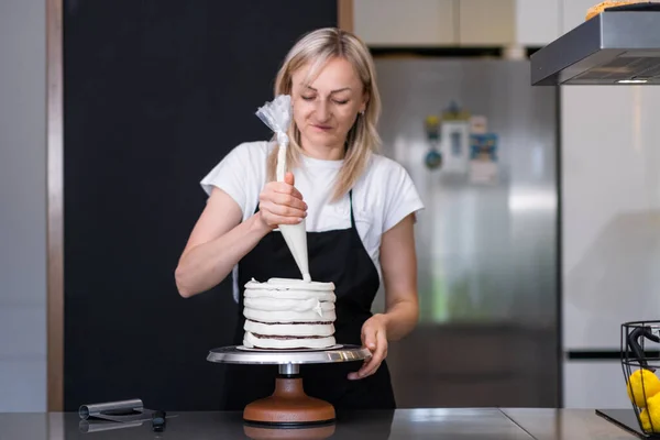 Focused food blogger showing process of making creamy homemade cake to followers on social media blonde woman in apron putting white mastic on baked dough with pastry cone
