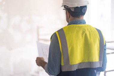 Profesyonel mühendis, eski püskü duvarlara bakıyor. Tadilat projesini elinde tutuyor. Kasketli ve yelekli adam odada yeniden yapılanma sürecine hazırlanıyor.