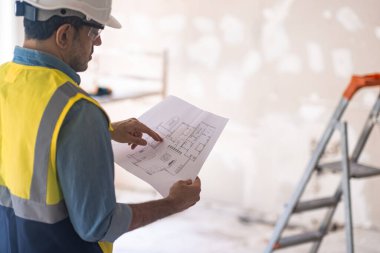 Konsantre işçiler miğferlerini kontrol ediyor. Daire çizimleri odada duruyor. Üniformalı bir uzman, eski püskü bir duvarın yanında merdivene karşı duruyor.