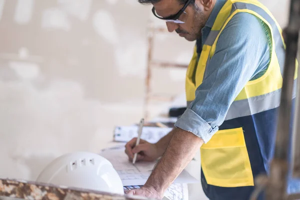 stock image Focused architect writing with ball-pen edits renovation project documentation specialist in uniform standing near construction safety equipment in unfinished premise