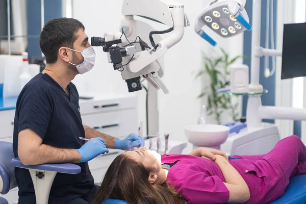 Diş mikroskobuyla genç bir kadının dişlerini kontrol etmek için profesyonel ekipmanlarla maske takan kadın dişçi muayenesi.