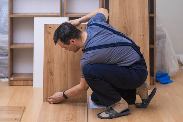 Custom furniture production and delivery man taking wooden planks for shelves to attach to cabinet near sofa under film firm worker in professional uniform
