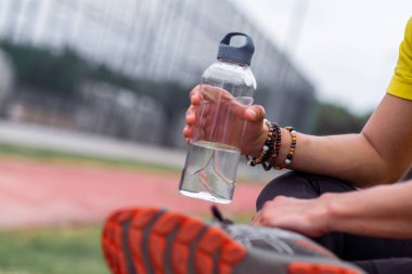 Spor salonundaki antrenmandan sonra elinde plastik su şişesi tutan boncuk bilezikli sporcu şehir stadyumunda içecekle oturuyor.