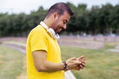 Yeşil şehir parkında akıllı telefona bakarken gülümseyen, şehir spor salonunda cep telefonu kullanan sarı tişörtlü bir adam.