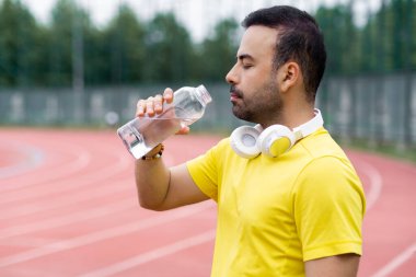 Telsiz kulaklıklı bir adam açık su şişesine bakıyor ve spor sahasında antrenman yaptıktan sonra dinleniyor. Sakallı sporcu içkiyle ferahlatıyor.