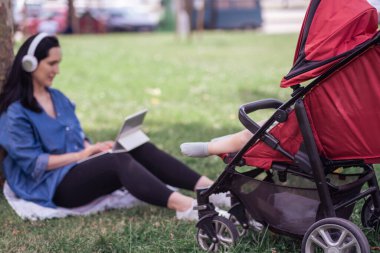 Kulaklıklı bir kadın, yeşil parktaki çimlerde oturan tabletle internetten bilgi öğreniyor. Bebek arabasının gölgesinde rüya gören sakin küçük bebeğe odaklanıyor.