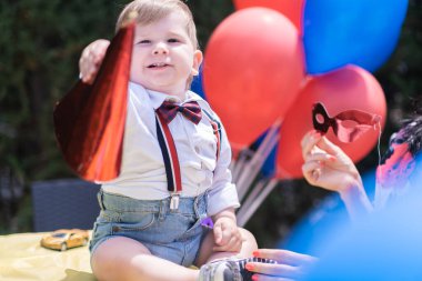 İlk doğum günü şenlikli bir bebek, Mutlu yıllar şapkalı, açık hava partisi kırmızı ve mavi balonlarla süslenmiş.