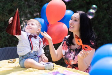 Gülümseyen bebek özel günü - 1 yo bebek mutluluğu dışarıda ilk doğum gününü kutluyor, kırmızı ve mavi balonlarla çevrili, kabarcıklarla meşgul olan ve sabun köpükleri kovalayan. 