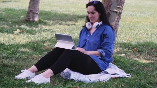 ハッピーブルネット女性とともにコードレスヘッドフォン保持タブレットコンピュータ座っていますによって木で緑の夏の庭フリーランサー仕事仕事をするインターネット屋外 — ストック動画