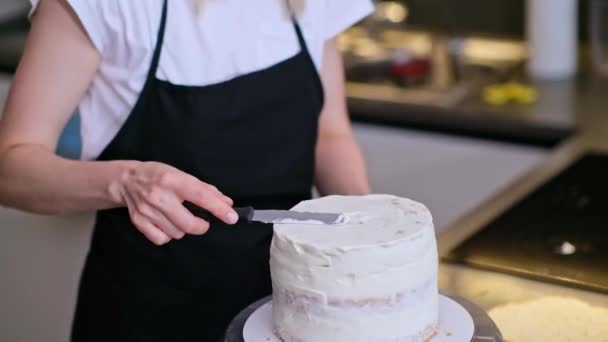 全神贯注的金发女人在烤蛋糕周围涂着白霜 站在厨房里 穿着黑色围裙的自制糖果商 在家里享受着小小的糖果生意 — 图库视频影像