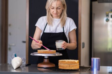 Siyah önlüklü konsantre şekerleme. Pasta tabaklarını taze kremayla süpürüyor. Mutfak masasında duran fırça sarışını kullanarak lezzetli şekerlemeler yapıyor.